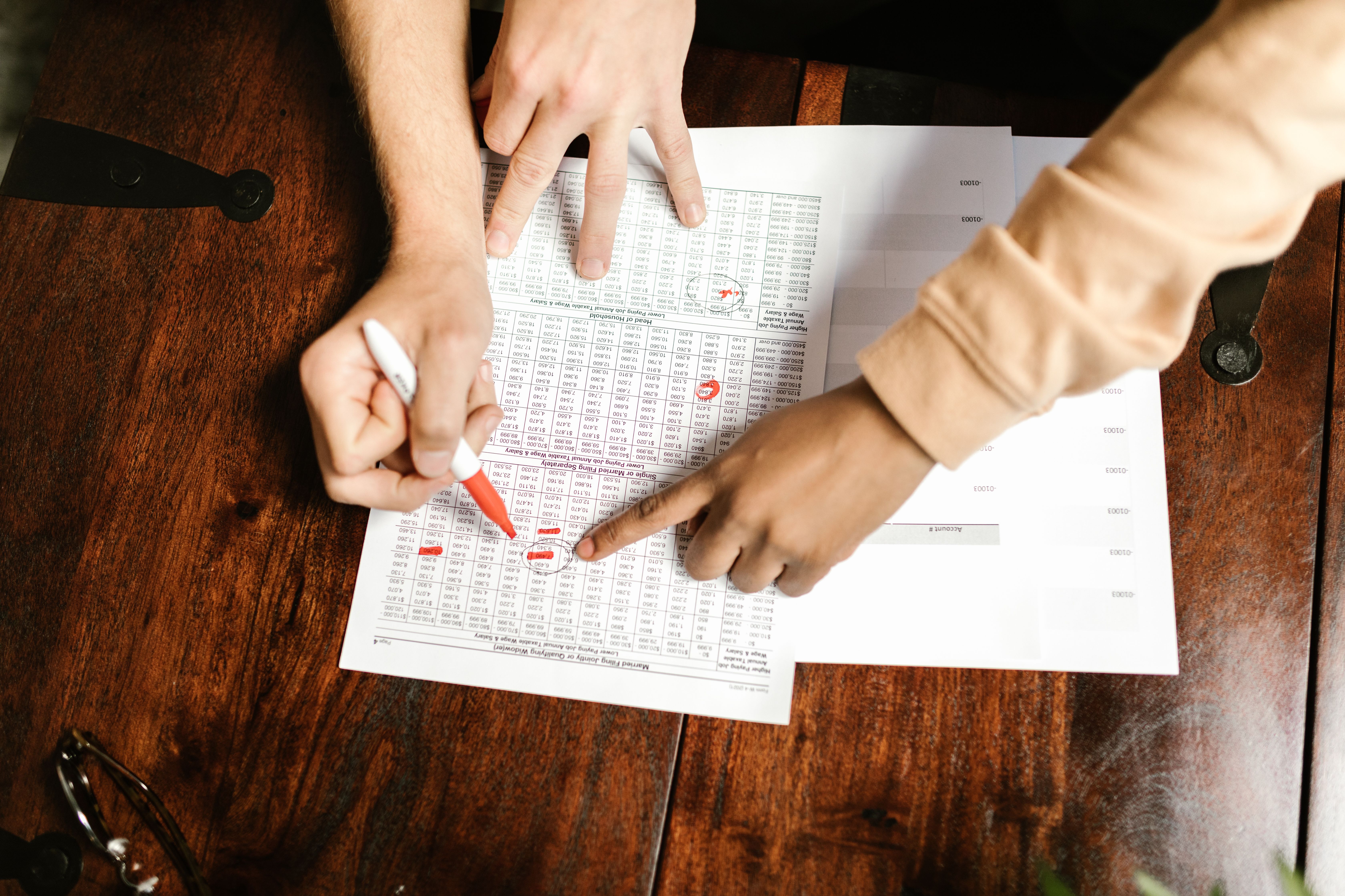 Two hands reviewing and marking financial documents with a red pen on a wooden table